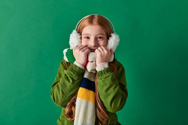 Menina bonito em cachecol listrado e abafadores de ouvido de inverno sorrindo em pano de fundo turquesa, camadas aconchegantes — Fotografia de Stock