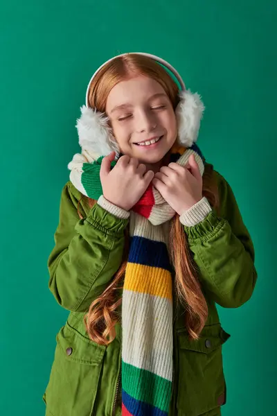 Menina satisfeito em cachecol listrado e abafadores de ouvido de inverno sorrindo em pano de fundo turquesa, camadas aconchegantes — Fotografia de Stock