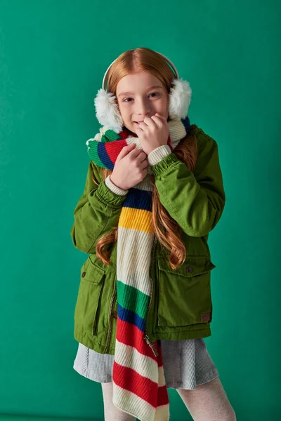 Chica alegre en bufanda rayada y orejeras de invierno sonriendo sobre fondo turquesa, capas acogedoras - foto de stock