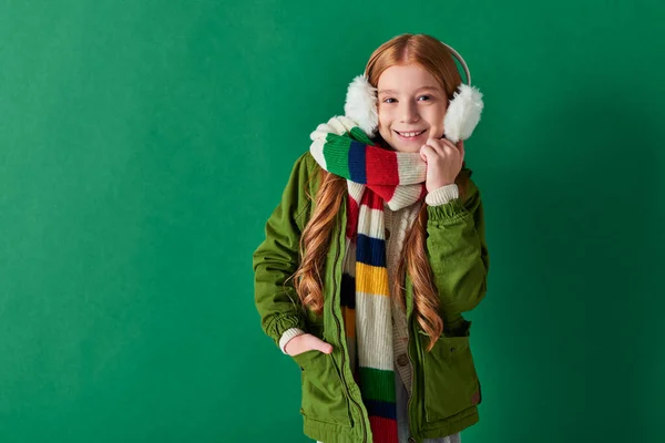 Alegre niña preadolescente en orejeras, bufanda rayada y traje de invierno posando sobre fondo turquesa - foto de stock