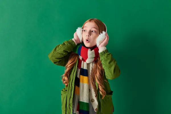 Шокированная девочка-подросток в наушниках, полосатый шарф и зимний наряд, смотрящая на бирюзовый — стоковое фото