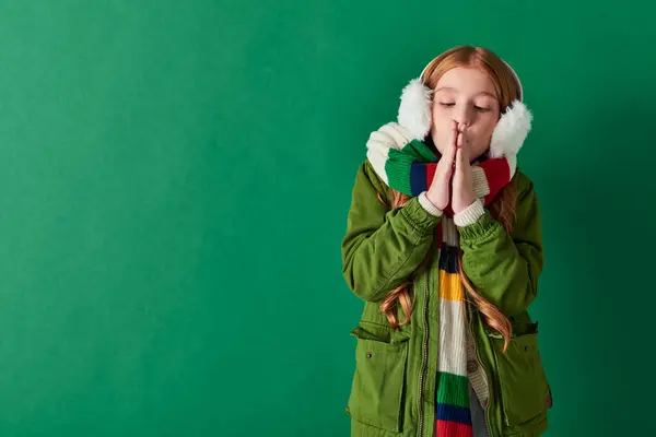 Preteen girl in ear muffs, striped scarf and winter outfit warming up hands on turquoise backdrop — Stock Photo