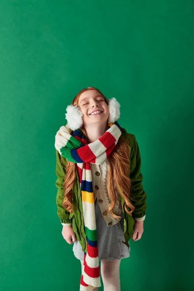 Happy preteen girl in ear muffs, striped scarf and winter outfit standing on turquoise backdrop — Stock Photo