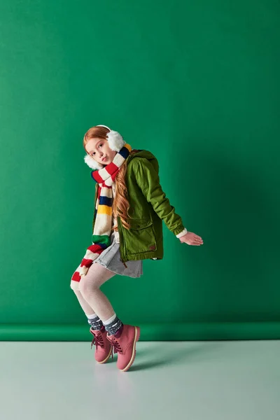 Stylisches Preteen Girl in Ohrenschützern, gestreiftem Schal und Winteroutfit posiert vor türkisfarbenem Hintergrund — Stockfoto