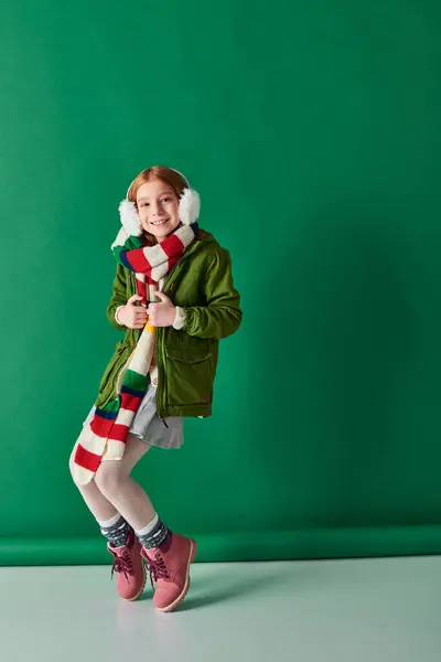 Весела дівчина на вушних муфтах, смугастий шарф і зимовий одяг позує на бірюзовому тлі — стокове фото