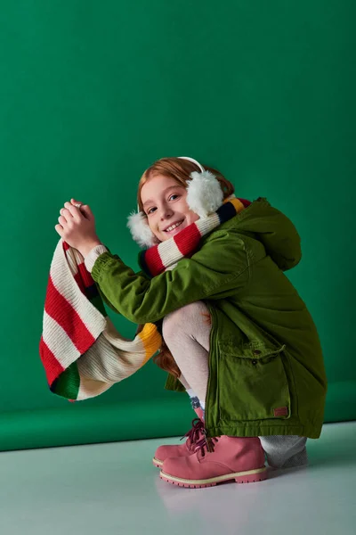 Menina pré-adolescente alegre em regalos auriculares, cachecol listrado e roupa de inverno sentado em pano de fundo turquesa — Fotografia de Stock