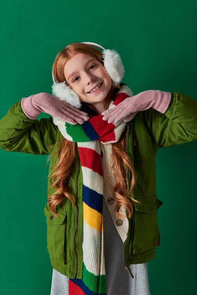 Happy preteen girl in ear muffs, striped scarf and winter outfit smiling on turquoise backdrop — Stock Photo