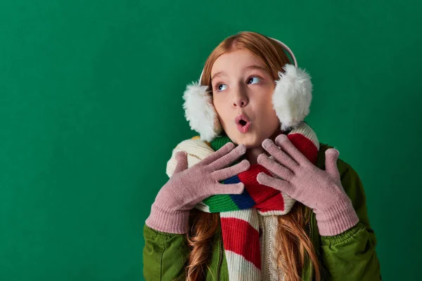 Schockiertes Mädchen in Ohrenschützern, gestreiftem Schal und Winteroutfit gestikuliert vor türkisfarbenem Hintergrund — Stockfoto