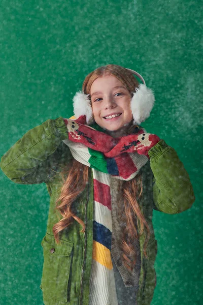 Positives Kind mit Ohrenschützer, gestreiftem Schal und Winterkleidung, das unter fallendem Schnee auf Türkis steht — Stockfoto