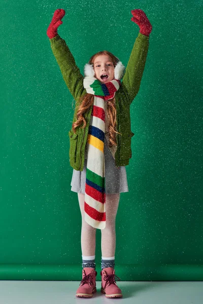Menina animado em muffs ouvido, cachecol listrado e traje de inverno de pé sob a neve caindo em turquesa — Fotografia de Stock