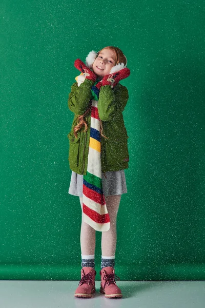 Chica feliz en orejeras, bufanda caliente y atuendo de invierno de pie bajo la nieve que cae en turquesa - foto de stock