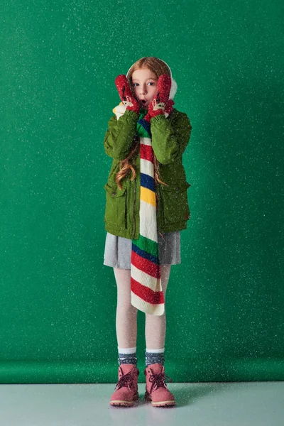 Überraschtes Mädchen in Ohrenschützer, Schal und Winterkleidung steht unter fallendem Schnee auf Türkis — Stockfoto