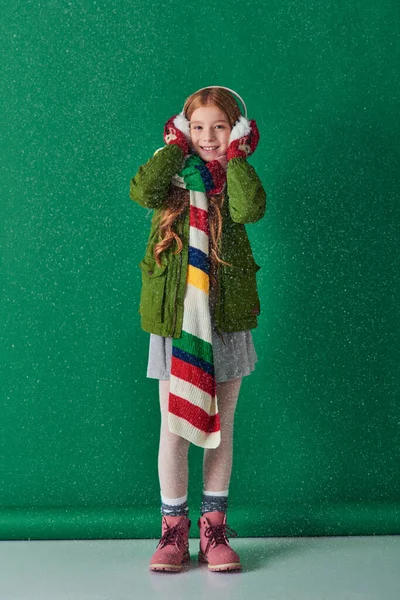 Fröhliches Mädchen in Ohrenschützern, warmem Schal und Winterkleidung, das unter fallendem Schnee auf Türkis steht — Stockfoto