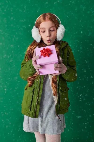 Preteen girl in ear muffs, scarf and winter attire blowing snow from Christmas present on turquoise — Stock Photo