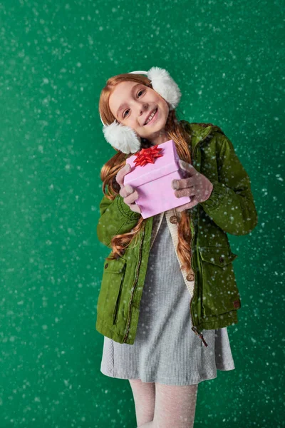 Pleased girl in ear muffs, scarf and winter attire holding Christmas present under falling snow — Stock Photo