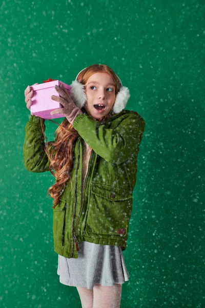 Stupito ragazza in cuffie, sciarpa e abbigliamento invernale tenendo regalo di Natale sotto la neve caduta — Foto stock