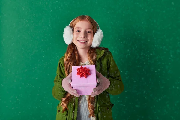Season of joy, happy kid in ear muffs and winter outfit holding Christmas gift under falling snow — Stock Photo