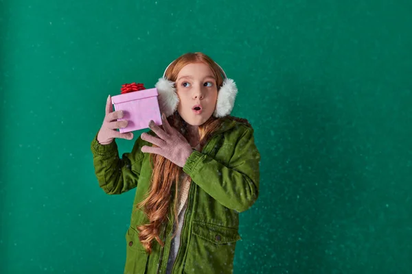 Curious girl in ear muffs, scarf and winter attire holding Christmas present under falling snow — Stock Photo