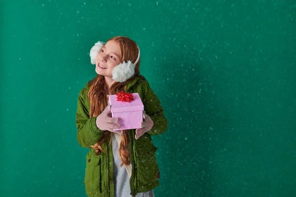 Temporada de alegría, complacido niño con orejeras y traje de invierno celebración de regalo de Navidad bajo la nieve caída - foto de stock