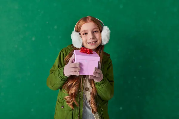 Temporada de alegria, criança feliz em regalos auriculares e roupa de inverno segurando presente de férias sob queda de neve — Fotografia de Stock
