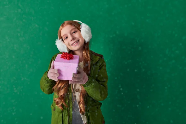 Season of joy, cheerful kid in ear muffs and winter outfit holding holiday gift under falling snow — Stock Photo