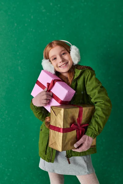 Season of joy, happy kid in ear muffs and winter outfit holding holiday gifts under falling snow — Stock Photo