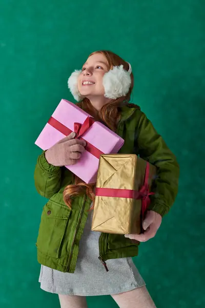 Season of joy, pleased girl in winter outfit and ear muffs holding holiday gifts under falling snow — Stock Photo