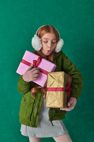 Season of joy, excited girl in winter outfit and ear muffs holding holiday gifts under falling snow — Stock Photo