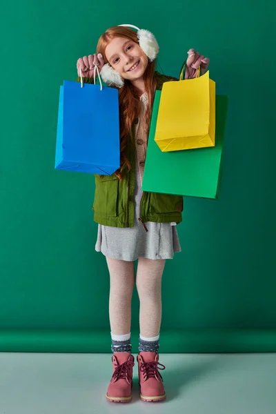 Preto sexta-feira e feriado temporada, criança feliz em roupas de inverno e abafadores de ouvido segurando sacos de compras — Fotografia de Stock