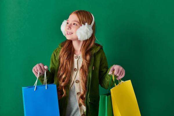 Holiday season, happy girl in winter outfit and ear muffs holding shopping bags on turquoise — Stock Photo