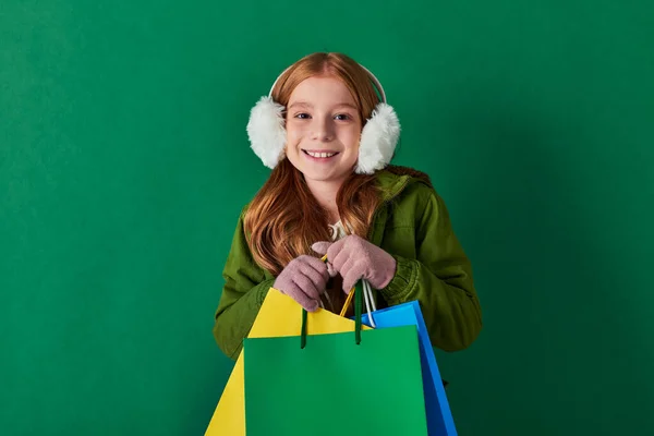 Temporada de férias, garoto animado em roupas de inverno e abafadores de ouvido segurando sacos de compras em turquesa — Fotografia de Stock