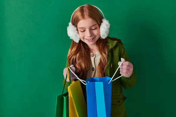 Зимний отдых, счастливый ребенок в зимнем наряде и наушники глядя внутрь сумки с покупками на бирюзовый — стоковое фото
