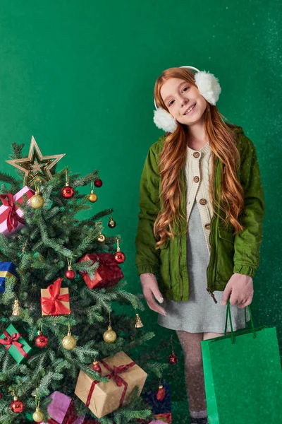 Vacaciones de invierno, chica feliz en orejeras sosteniendo bolsas de compras cerca del árbol de Navidad, nieve cayendo - foto de stock