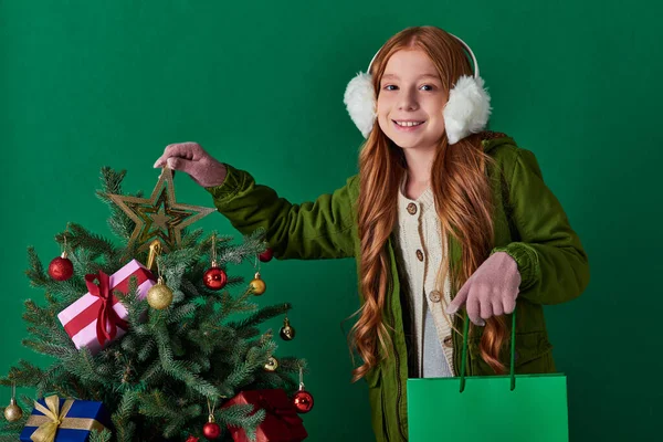 Vacaciones de invierno, chica feliz en orejeras sosteniendo bolsa de compras tocando la parte superior de la estrella del árbol de Navidad - foto de stock