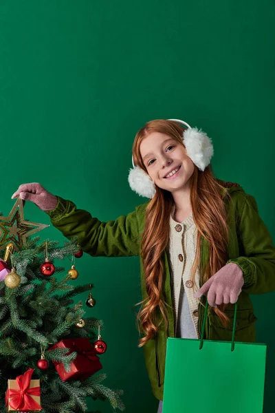 Winter holidays, cheerful girl in ear muffs holding shopping bag touching star top of Christmas tree — Stock Photo