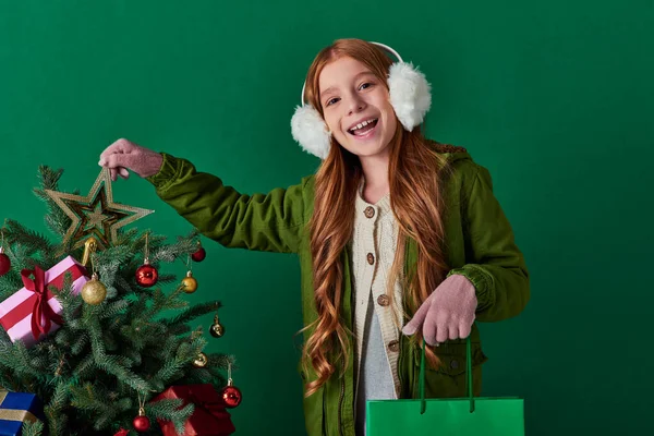 Winter holidays, excited girl in ear muffs holding shopping bag touching star top of Christmas tree — Stock Photo