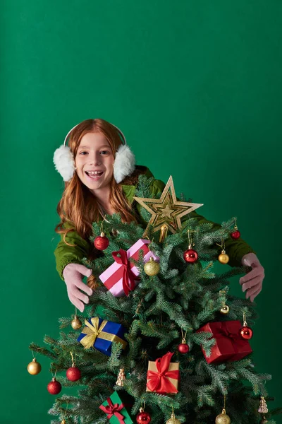 Holidays, happy girl in ear muffs standing behind decorated Christmas tree on turquoise backdrop — Stock Photo