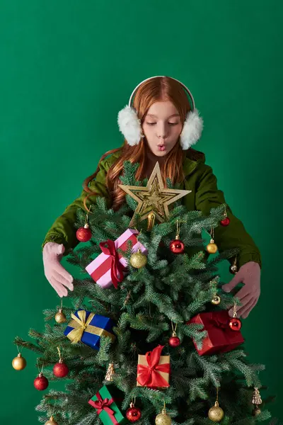 Holidays, amazed girl in ear muffs standing behind decorated Christmas tree on turquoise backdrop — Stock Photo