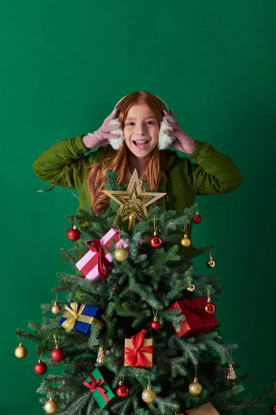 Vacanze, ragazza eccitata indossa manicotti per le orecchie e in piedi dietro l'albero di Natale decorato su turchese — Foto stock