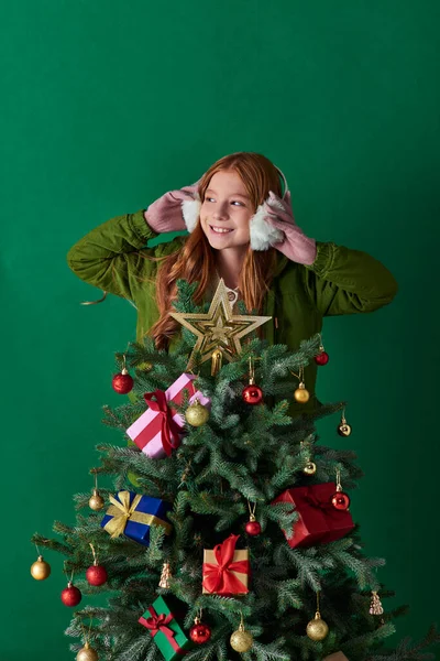 Holidays, cheerful girl wearing ear muffs and standing behind decorated Christmas tree on turquoise — Stock Photo