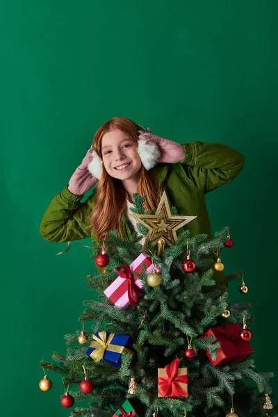Esprit de vacances, fille heureuse portant des cache-oreilles et debout près décoré arbre de Noël sur turquoise — Photo de stock