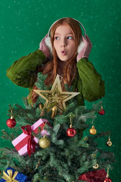 Holiday spirit, surprised girl in ear muffs hugging decorated Christmas tree on turquoise backdrop — Stock Photo