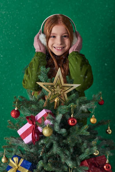 Holiday joy, happy girl in ear muffs hugging decorated Christmas tree on turquoise backdrop — Stock Photo