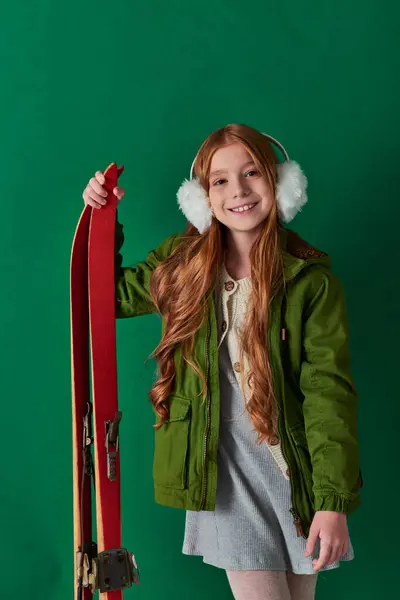 Niña preadolescente feliz en orejeras y traje de invierno sonriendo y sosteniendo esquís rojos sobre fondo turquesa - foto de stock
