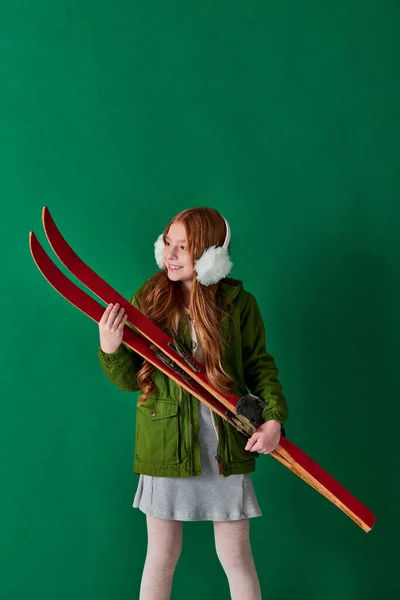 Alegre niña preadolescente en orejeras y traje de invierno sosteniendo equipo de esquí rojo sobre fondo turquesa - foto de stock