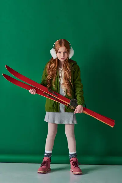 Angry preteen girl in ear muffs and winter outfit holding red ski gear on turquoise backdrop — Stock Photo