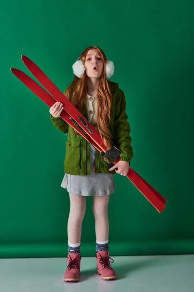 Cold air, preteen girl in ear muffs and winter outfit breathing and holding red skis on turquoise — Stock Photo