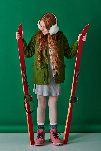 Cheerful preteen girl in ear muffs and winter outfit holding red skis on turquoise background — Stock Photo