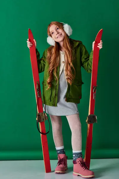 Excited preteen girl in ear muffs and winter attire holding red skis on turquoise background — Stock Photo