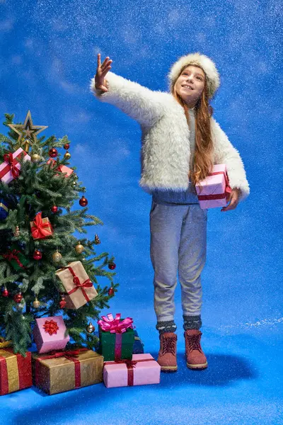 Cheerful girl in faux fur jacket and hat holding gift and catching snow near Christmas tree on blue — Stock Photo
